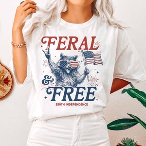 May include: White t-shirt with a graphic of a raccoon wearing sunglasses with the American flag design, holding a flag, and the words "FERAL & FREE" and "250th INDEPENDENCE" in red and blue.