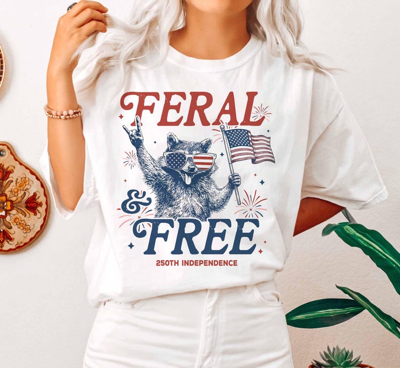 May include: White t-shirt with a graphic of a raccoon wearing sunglasses with the American flag design, holding a flag, and the words "FERAL & FREE" and "250th INDEPENDENCE" in red and blue.