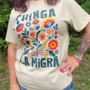 Op de afbeelding: Beige t-shirt met de tekst "CHINGA LA MIGRA" in teal. Het shirt heeft een kleurrijk bloemenontwerp met rode, oranje, gele en blauwe bloemen. Het shirt met korte mouwen is gemaakt van een zacht materiaal.