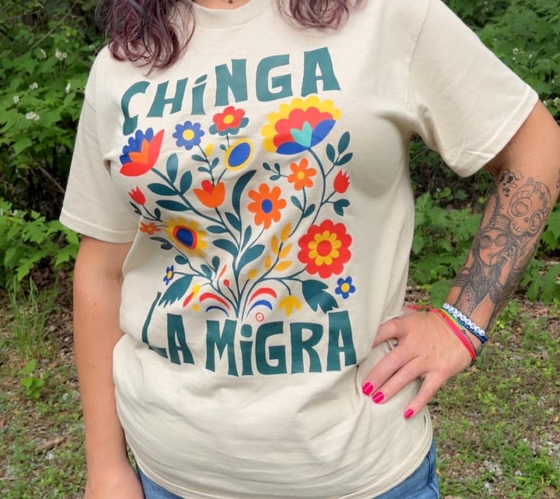 Op de afbeelding: Beige t-shirt met de tekst "CHINGA LA MIGRA" in teal. Het shirt heeft een kleurrijk bloemenontwerp met rode, oranje, gele en blauwe bloemen. Het shirt met korte mouwen is gemaakt van een zacht materiaal.