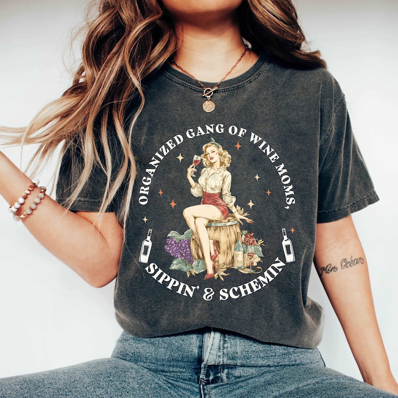 May include: Dark grey t-shirt with a circular graphic of a woman with wine, seated on a barrel. Text reads "ORGANIZED GANG OF WINE MOMS, SIPPIN' & SCHEMIN". Two wine bottles flank the graphic.