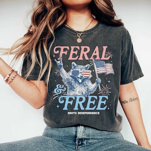 May include: Dark gray t-shirt with the words "FERAL & FREE" in pink and blue, featuring a raccoon wearing sunglasses with an American flag design. The raccoon is holding an American flag. The shirt also says "250TH INDEPENDENCE".