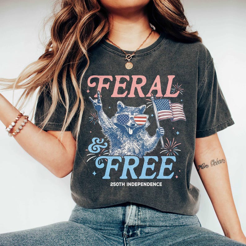 May include: Dark gray t-shirt with the words "FERAL & FREE" in pink and blue, featuring a raccoon wearing sunglasses with an American flag design. The raccoon is holding an American flag. The shirt also says "250TH INDEPENDENCE".
