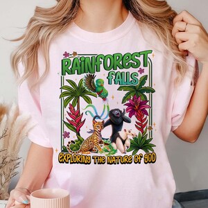 May include: Light pink t-shirt with a colorful rainforest graphic. The design features a parrot, a monkey, a frog, and other animals, along with the text "Rainforest Falls" and "Exploring the Nature of God."