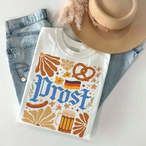 May include: White t-shirt with a colorful graphic design featuring the word "Prost" in blue, a pretzel, beer mug, and German flag. The shirt is laid on top of blue jeans and next to a tan hat.