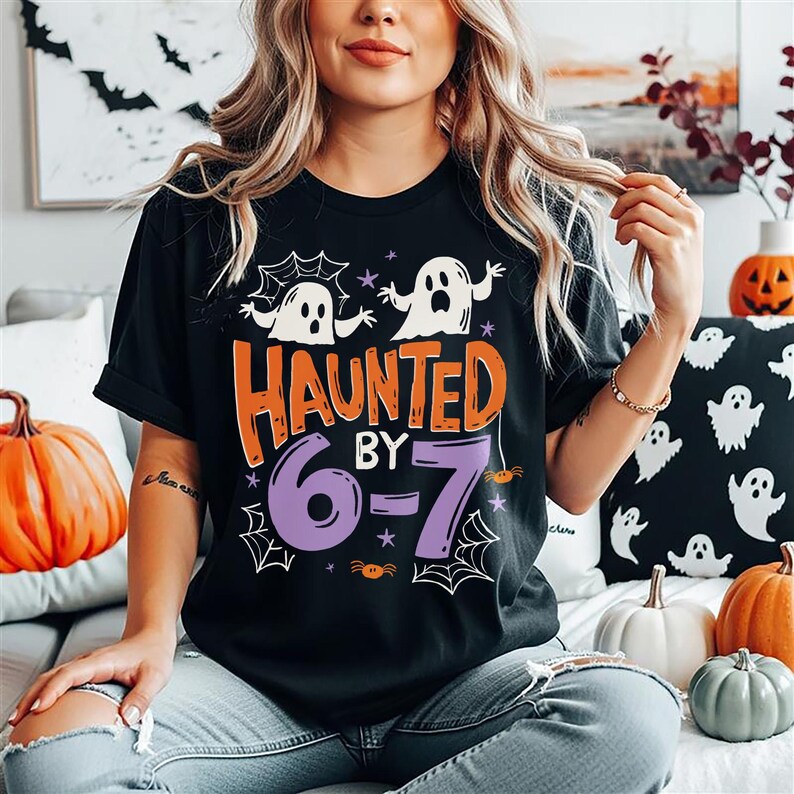 May include: Black t-shirt with the words "HAUNTED BY 6-7" in orange and purple, featuring ghost and spider web graphics. The shirt is part of a Halloween-themed collection.