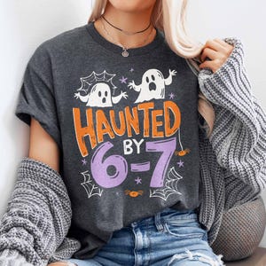 May include: Dark gray t-shirt with a Halloween-themed graphic. The design features two white ghosts, the words "HAUNTED BY 6-7" in orange and purple, spiderwebs, and small spiders. The shirt is a casual, short-sleeved style.