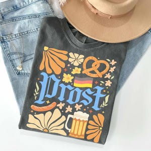 May include: A charcoal gray t-shirt with the word "Prost" in blue, featuring a pretzel, beer mug, sausage, and floral designs. A tan hat and light blue jeans are also in the image. A gold necklace is visible.