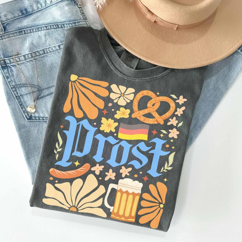 May include: A charcoal gray t-shirt with the word "Prost" in blue, featuring a pretzel, beer mug, sausage, and floral designs. A tan hat and light blue jeans are also in the image. A gold necklace is visible.