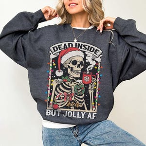 May include: A dark gray sweatshirt with a Christmas-themed graphic of a skeleton wearing a Santa hat and holding a mug. The text on the sweatshirt reads "Dead Inside But Jolly AF".