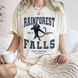 May include: Cream-colored t-shirt with the words "RAINFOREST FALLS" in navy blue, featuring a cartoon monkey. The text "DAY 5 HOWIE" is above the monkey, and "God is forever - Psalm 115:18" is below. The shirt has short sleeves.