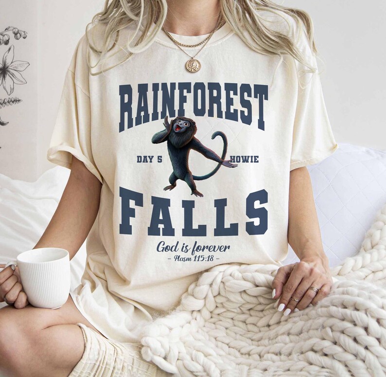 May include: Cream-colored t-shirt with the words "RAINFOREST FALLS" in navy blue, featuring a cartoon monkey. The text "DAY 5 HOWIE" is above the monkey, and "God is forever - Psalm 115:18" is below. The shirt has short sleeves.