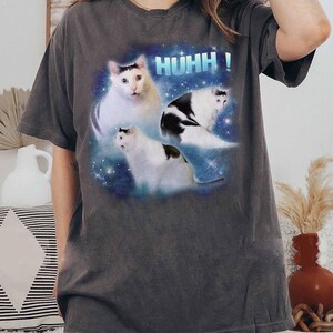 May include: Dark grey t-shirt with a graphic of three white cats with black markings against a blue and starry background. The word "HUHH!" is printed above the cats in blue block letters.