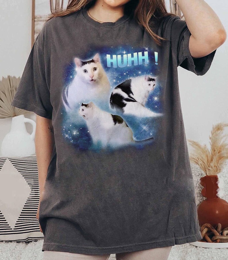 May include: Dark grey t-shirt with a graphic of three white cats with black markings against a blue and starry background. The word "HUHH!" is printed above the cats in blue block letters.
