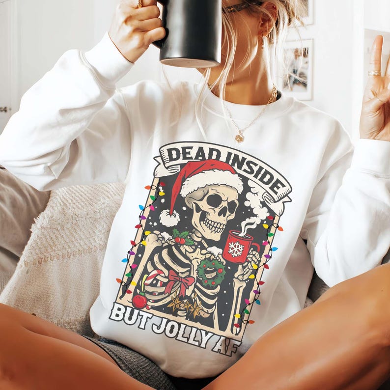 May include: White Christmas sweater with a skeleton wearing a Santa hat and holding a mug of hot cocoa. The sweater has the text "Dead Inside But Jolly AF" printed on it.