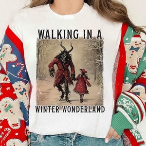 May include: White t-shirt featuring a winter-themed illustration. The graphic depicts a horned figure in a red coat holding hands with a child in a red dress, with the text "WALKING IN A WINTER WONDERLAND".