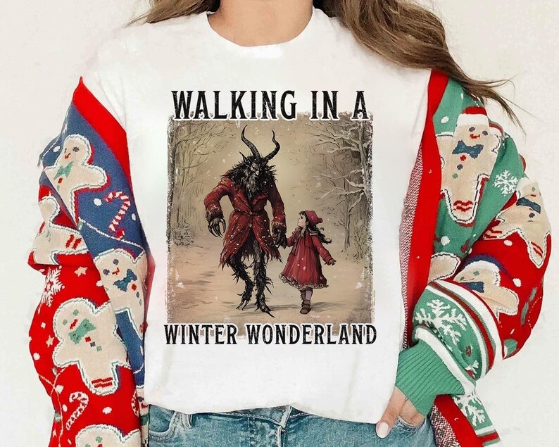 May include: White t-shirt featuring a winter-themed illustration. The graphic depicts a horned figure in a red coat holding hands with a child in a red dress, with the text "WALKING IN A WINTER WONDERLAND".