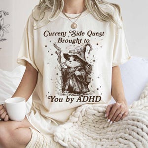 Pode incluir: Camiseta creme com uma ilustra&ccedil;&atilde;o de um gato mago segurando uma bengala. O texto diz "Current Side Quest Brought to You by ADHD". O design &eacute; em tons de marrom e preto.