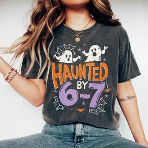 May include: Dark gray t-shirt with a Halloween-themed design. The shirt features the text "HAUNTED BY 6-7" in orange and purple, along with white ghost and spiderweb graphics. A fun and festive tee for the spooky season.
