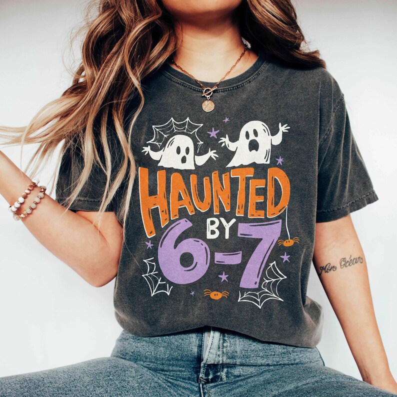 May include: Dark gray t-shirt with a Halloween-themed design. The shirt features the text "HAUNTED BY 6-7" in orange and purple, along with white ghost and spiderweb graphics. A fun and festive tee for the spooky season.