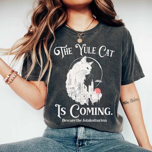May include: Dark gray t-shirt with the text "The Yule Cat Is Coming." A white cat illustration is centered on the shirt, set against a cityscape and a red sun. The phrase "Beware the Jolakottrinn" is printed below the main text.