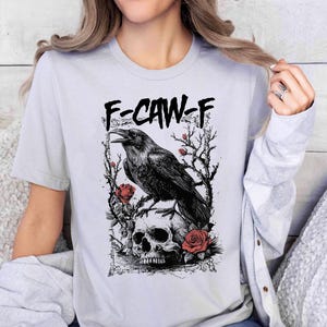 Puede incluir: Camiseta gris claro con un gr&aacute;fico negro de un cuervo posado sobre una calavera, rodeado de rosas y ramas. El texto "F-CAW-F" est&aacute; impreso encima del cuervo. El dise&ntilde;o es una ilustraci&oacute;n detallada.