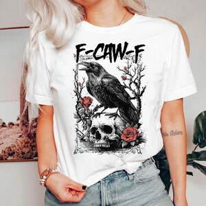 Puede incluir: Camiseta blanca con un dise&ntilde;o de cuervo negro posado sobre una calavera, rodeado de rosas y ramas espinosas. El texto "F-CAW-F" est&aacute; impreso encima del cuervo. Ilustraci&oacute;n g&oacute;tica.