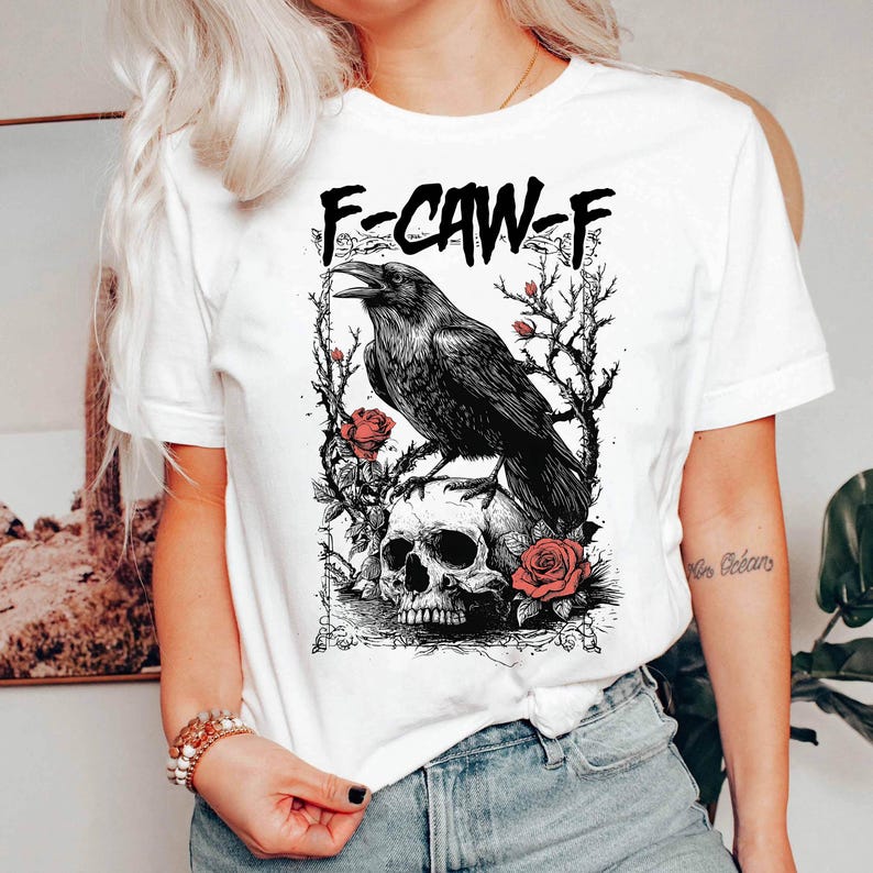 Puede incluir: Camiseta blanca con un dise&ntilde;o de cuervo negro posado sobre una calavera, rodeado de rosas y ramas espinosas. El texto "F-CAW-F" est&aacute; impreso encima del cuervo. Ilustraci&oacute;n g&oacute;tica.
