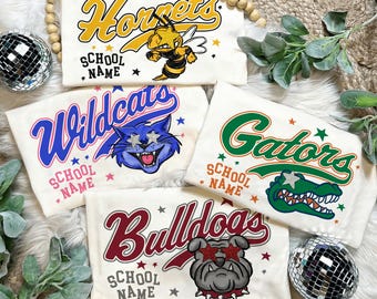 Może przedstawiać: Kremowe koszulki z wzorami maskotek szkolnych. Koszulki zawierają słowa "Hornets", "Wildcats", "Gators" i "Bulldogs", każda z grafiką maskotki i tekstem "School Name". Gwiazdy i elementy dekoracyjne otaczają tekst.