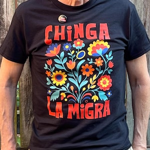 Op de afbeelding: Zwart t-shirt met de rode tekst "CHINGA LA MIGRA" boven een kleurrijk bloemenpatroon. De bloemen zijn oranje, geel, blauw en rood. Een kleine button is vastgespeld bij de halslijn.