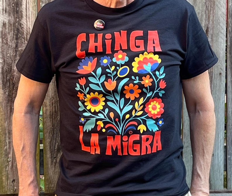 Op de afbeelding: Zwart t-shirt met de rode tekst "CHINGA LA MIGRA" boven een kleurrijk bloemenpatroon. De bloemen zijn oranje, geel, blauw en rood. Een kleine button is vastgespeld bij de halslijn.
