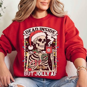 May include: Red Christmas sweater with a skeleton wearing a Santa hat and holding a mug of hot cocoa. The sweater has the text "Dead Inside But Jolly AF" printed on it.