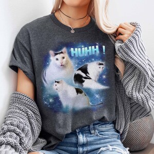 May include: Dark grey t-shirt with a graphic of three cats against a space-themed backdrop and the word "HUHH!". The cats are white with black markings. The shirt is made of a soft material.