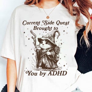 Pode incluir: Camiseta branca com uma ilustra&ccedil;&atilde;o de um gato vestido de mago, segurando uma bengala. O texto diz "Current Side Quest Brought to You by ADHD". O design &eacute; em tons de marrom e preto.