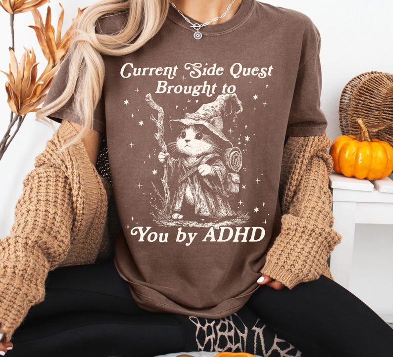 Pode incluir: Camiseta marrom com o texto "Current Side Quest Brought to You by ADHD". O gr&aacute;fico apresenta um gato vestido de mago com um bast&atilde;o e uma mochila. A camiseta tem um corte folgado.