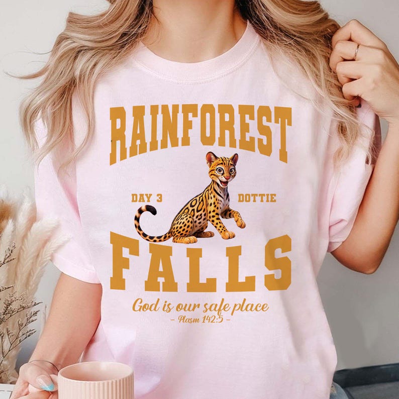 May include: Light pink t-shirt with a graphic featuring the text "RAINFOREST FALLS" in gold block letters, with a cartoon ocelot illustration. The text "Day 3 Dottie" and "God is our safe place - Psalm 142:5" are also included.