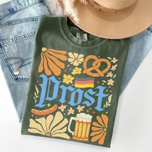May include: An olive green t-shirt with a colorful graphic design featuring the word "Prost" in blue, a pretzel, beer mug, sausage, and flowers. The shirt is laid on top of blue jeans and a tan hat.