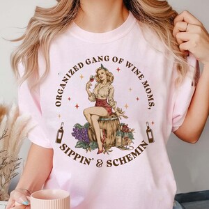 May include: Pale pink t-shirt with a graphic. It shows a vintage-style illustration of a woman on a wine barrel, holding a glass of wine. The text reads "ORGANIZED GANG OF WINE MOMS, SIPPIN' & SCHEMIN".