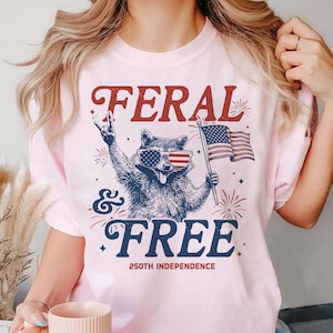 May include: Light pink t-shirt with a graphic of a raccoon wearing American flag sunglasses and holding an American flag. The text reads "FERAL & FREE" with "250TH INDEPENDENCE" below.