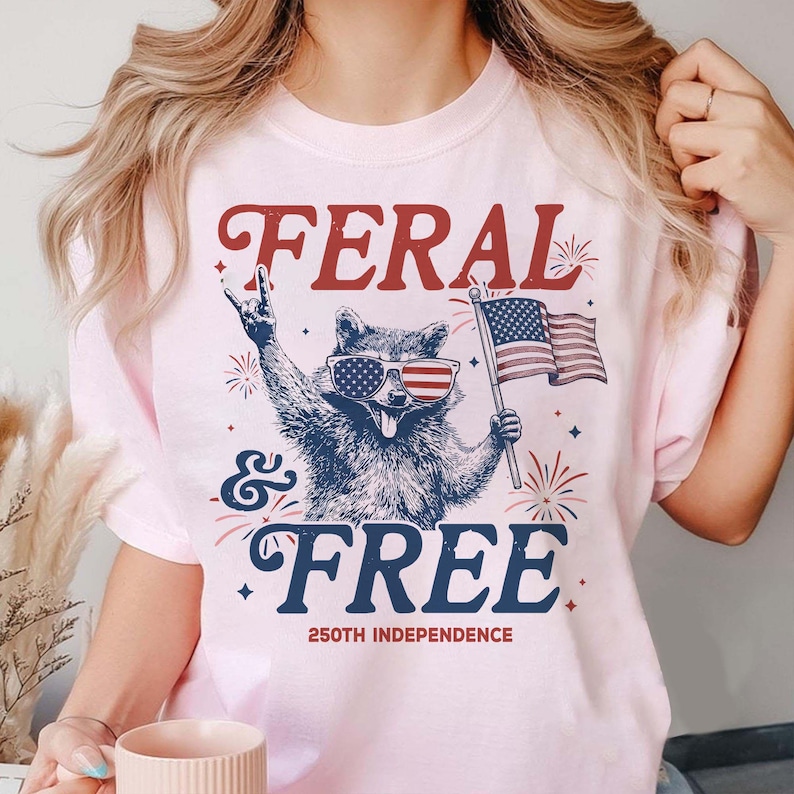 May include: Light pink t-shirt with a graphic of a raccoon wearing American flag sunglasses and holding an American flag. The text reads "FERAL & FREE" with "250TH INDEPENDENCE" below.