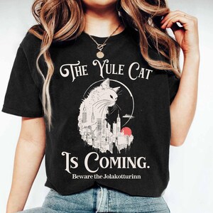 May include: Black t-shirt featuring a white cat illustration with a cityscape design inside a circle. The text "The Yule Cat Is Coming" is above and "Beware the Jolakotturinn" is below the cat graphic.