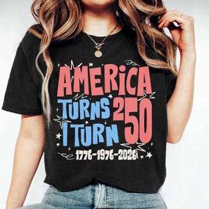 May include: Black t-shirt with the text "AMERICA TURNS 250 I TURN 50" in red and blue, with the years 1776-1976-2026 in white. The shirt has a crew neck and short sleeves.