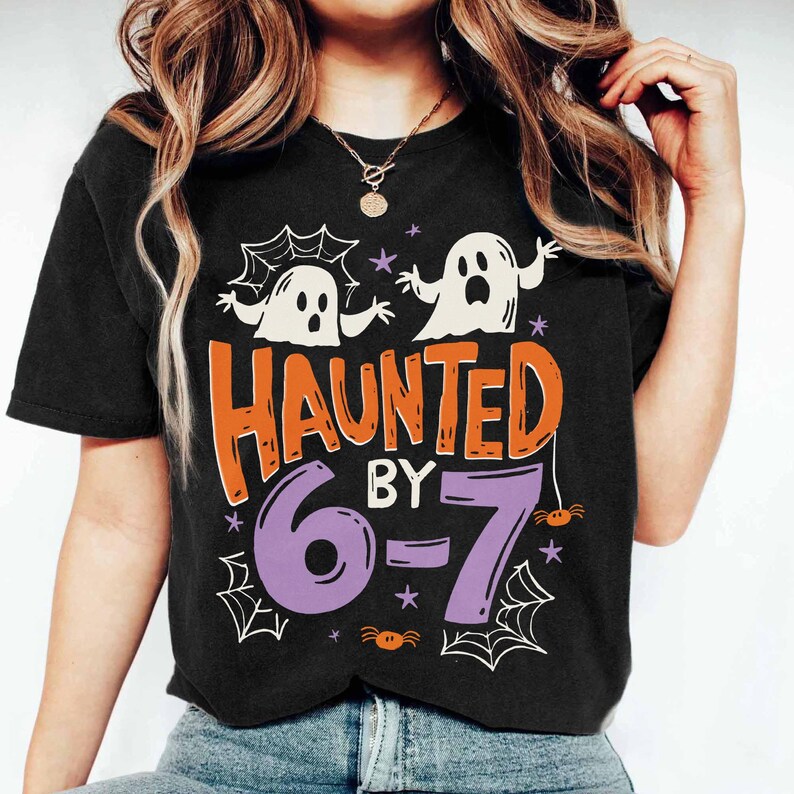 May include: Black t-shirt with a Halloween-themed graphic. The design features two white ghost figures, the words "HAUNTED BY" in orange, and the numbers "6-7" in purple. Spiderwebs and stars complete the design.