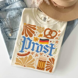 May include: Cream-colored t-shirt with a retro-style design featuring the word "Prost" in blue, a pretzel, a beer stein, and the German flag. The shirt also has floral and sausage graphics.