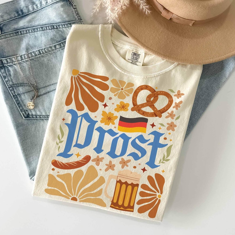 May include: Cream-colored t-shirt with a retro-style design featuring the word "Prost" in blue, a pretzel, a beer stein, and the German flag. The shirt also has floral and sausage graphics.