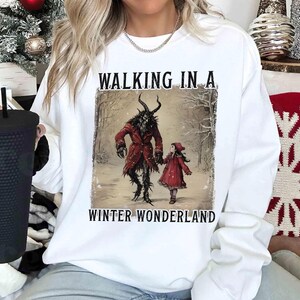 May include: White sweatshirt featuring a graphic of a horned figure in a red coat and a girl in a red dress, walking in a snowy forest. The text "WALKING IN A WINTER WONDERLAND" is above and below the image.