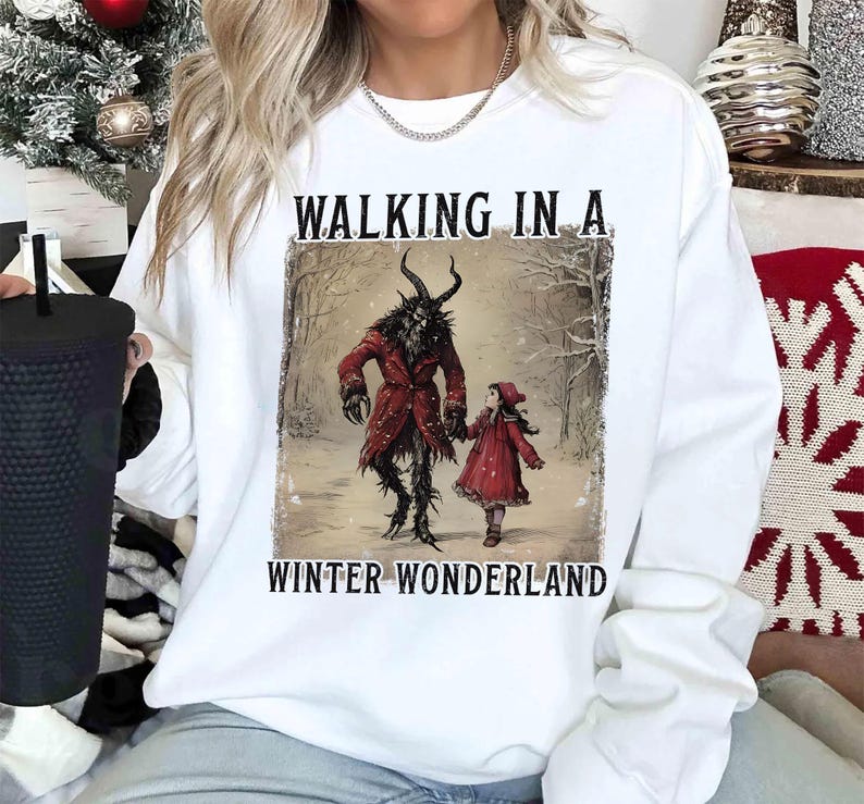 May include: White sweatshirt featuring a graphic of a horned figure in a red coat and a girl in a red dress, walking in a snowy forest. The text "WALKING IN A WINTER WONDERLAND" is above and below the image.