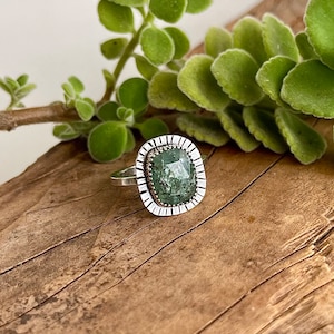 May include: A silver ring featuring a large, rectangular green gemstone with a textured surface. The gemstone is set within a decorative, scalloped bezel. The ring is displayed on a wooden surface, with green foliage in the background.