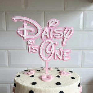 Puede incluir: Una tarta redonda de dos pisos con glaseado blanco, lunares negros y adornos de lazo de Minnie Mouse rosa. Un adorno para tarta rosa y blanco que dice "Daisy is One" en una fuente inspirada en Disney. Una tarta festiva para una celebración de primer cumpleaños.