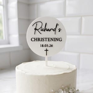 以下が含まれることがあります： 白いフロストケーキ、白い花と丸いケーキトッパーで飾られています。トッパーには「Richard's CHRISTENING 18.03.26」と十字架が書かれています。ケーキは白いケーキスタンドの上にあります。